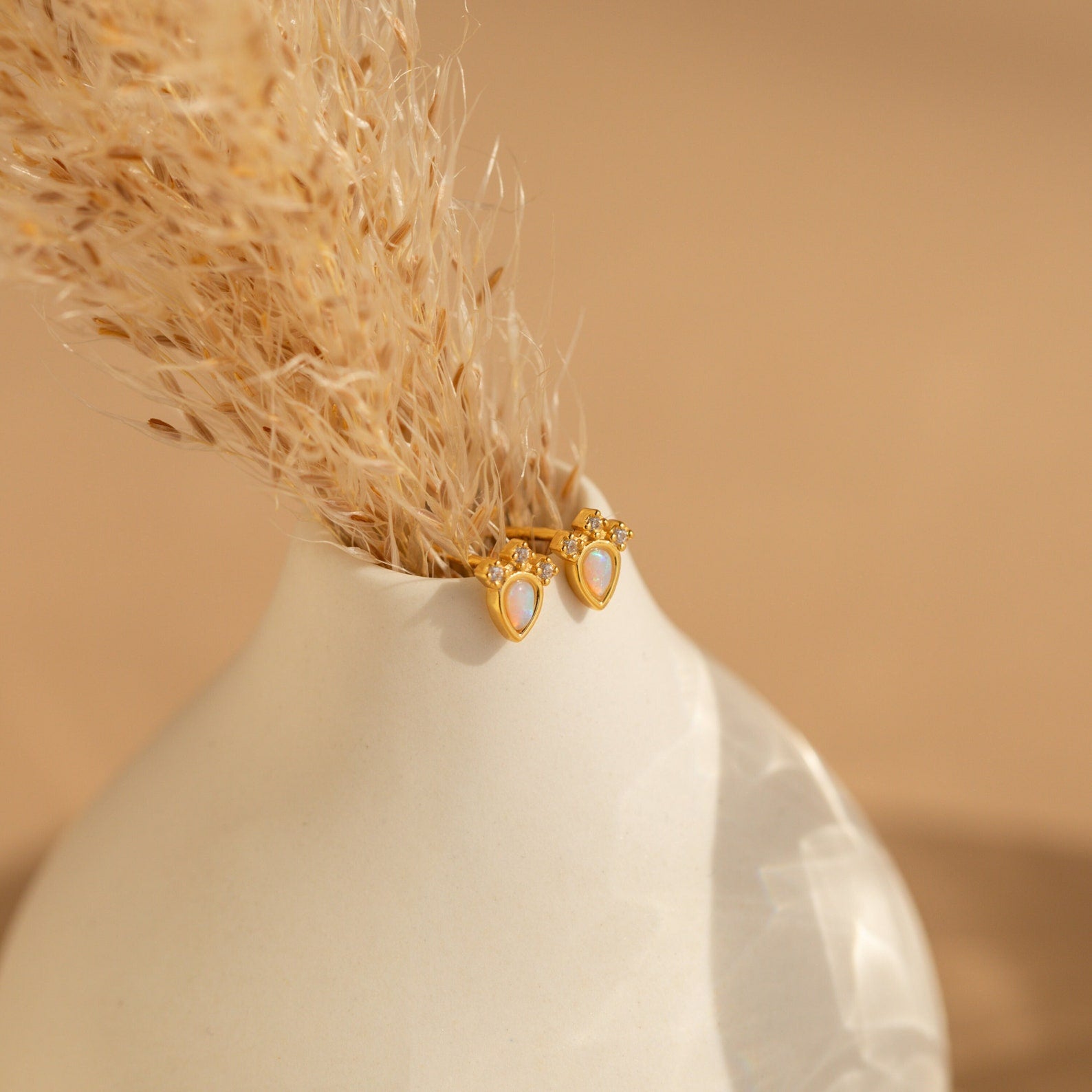 The Dainty Opal Teardrop Studs are displayed on a white vase with dried grass, set against a neutral background.