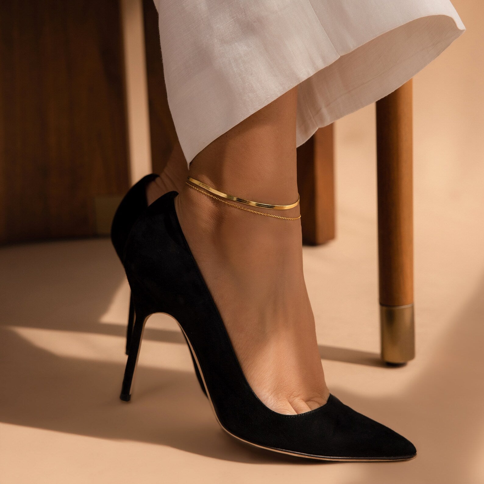 A person in white pants and black heels, seated on a wooden chair, models the Micro Rolo Chain Anklet by Caitlyn Minimalist.