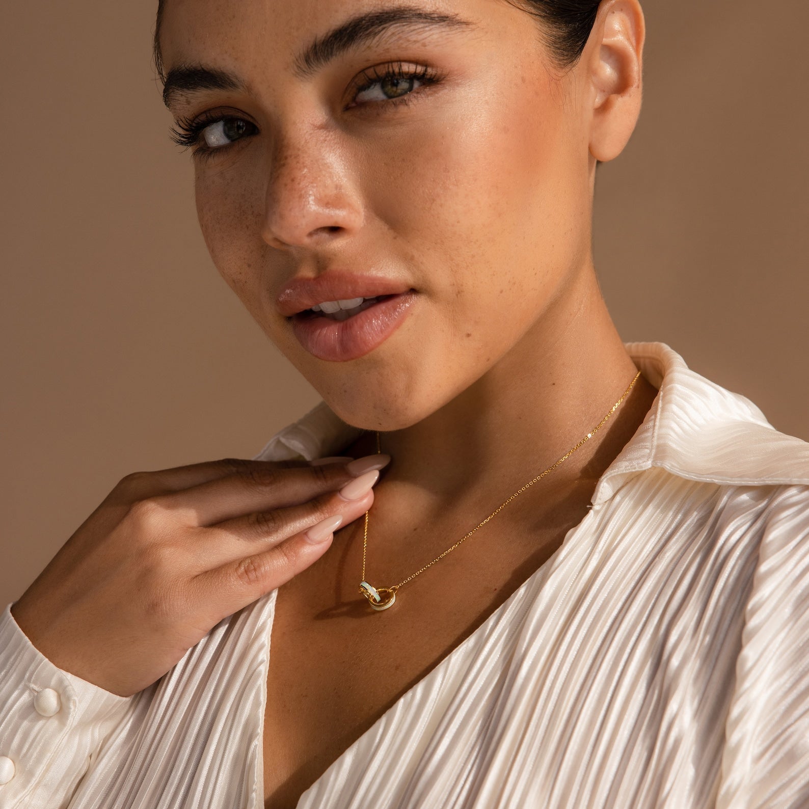 A woman in a white blouse touches the Interlocking Opal Inlay Necklace, symbolizing infinite love, while looking at the camera with a neutral background—an ideal anniversary gift.