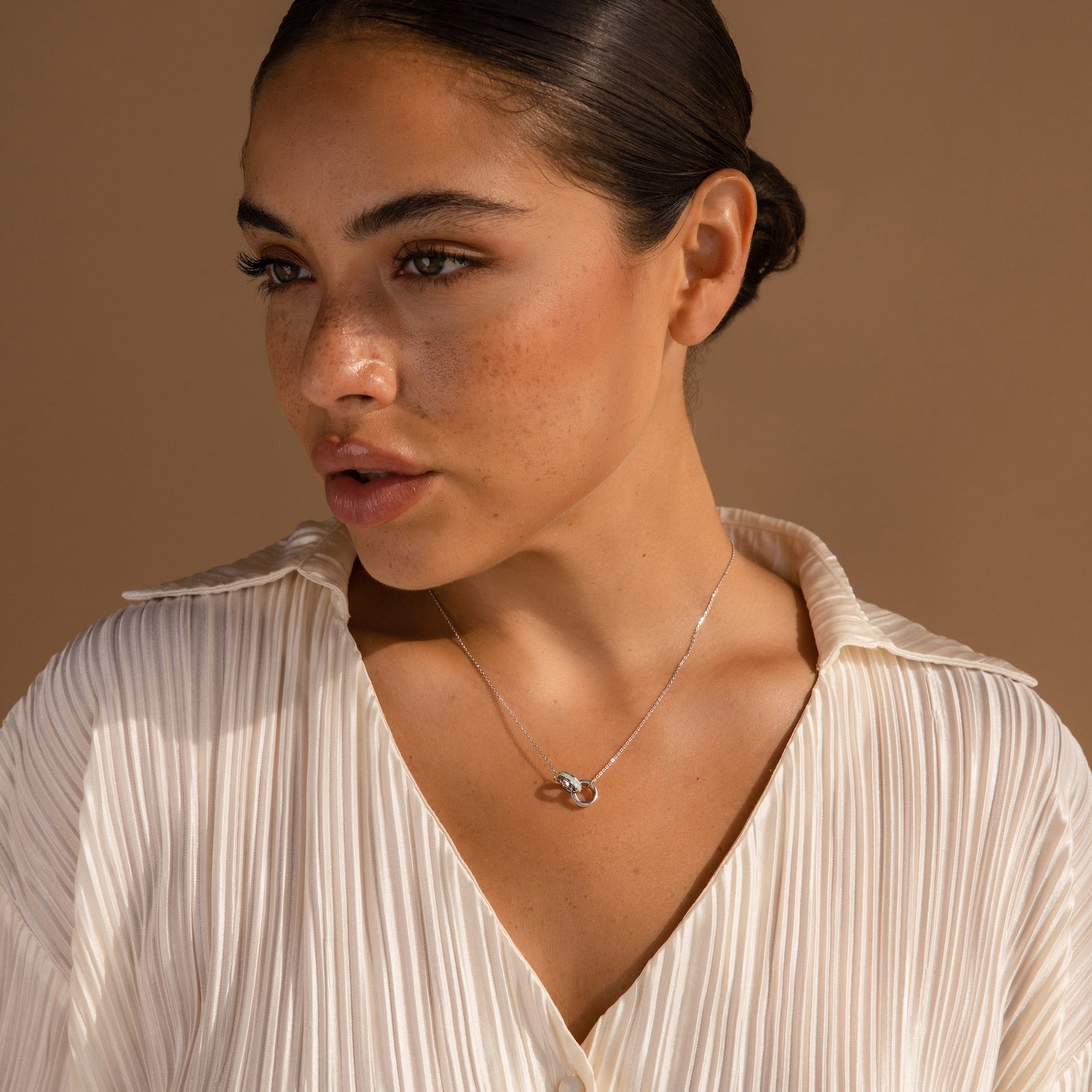 A woman with slicked-back hair wears a cream, pleated blouse and the Interlocking Opal Inlay Necklace—symbolizing infinite love—against a light brown background. An ideal anniversary gift.