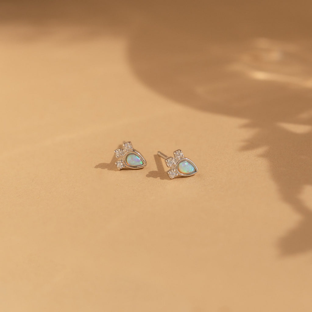 The Dainty Opal Teardrop Studs are showcased on a soft beige surface, their silver setting and teardrop shape highlighted by gentle shadows.