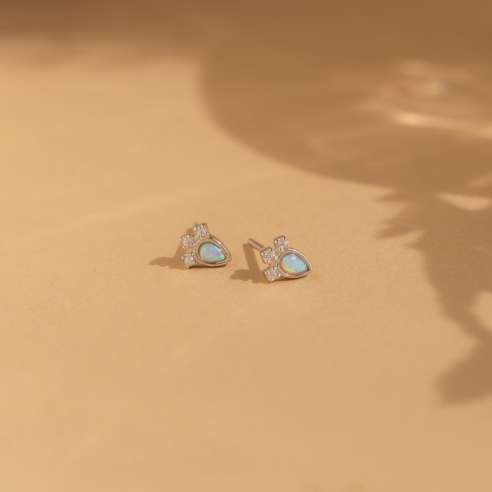 The Dainty Opal Teardrop Studs are showcased on a soft beige surface, their silver setting and teardrop shape highlighted by gentle shadows.