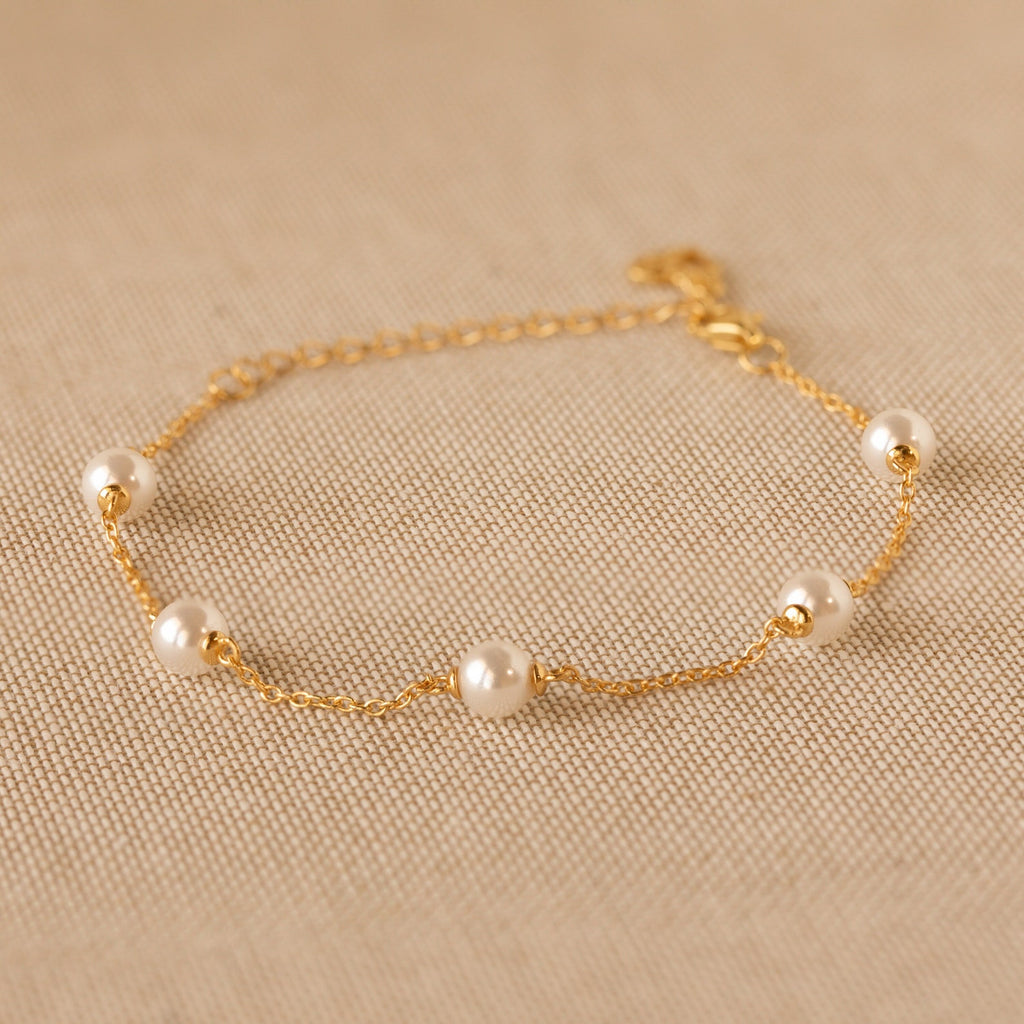 The Pearl Station Bracelet features five evenly spaced pearls on a delicate gold chain, elegantly displayed on beige textured fabric—a timeless piece of modern jewelry.