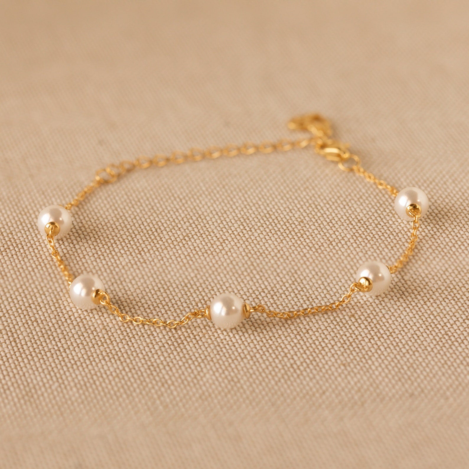 The Pearl Station Bracelet features five evenly spaced pearls on a delicate gold chain, elegantly displayed on beige textured fabric—a timeless piece of modern jewelry.