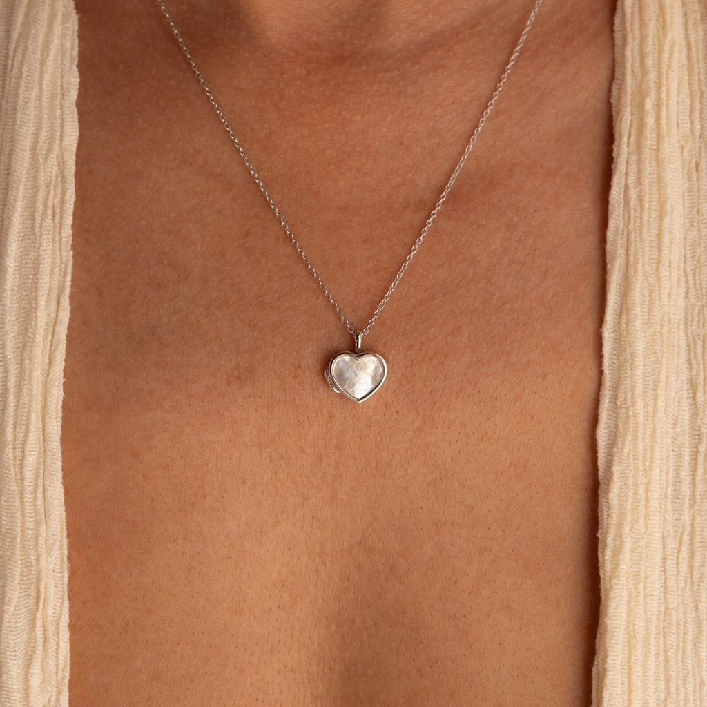 The Pearl Heart Locket Necklace, a nostalgic silver pendant, rests on the bare chest and is paired with a cream-colored garment.