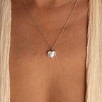 The Pearl Heart Locket Necklace, a nostalgic silver pendant, rests on the bare chest and is paired with a cream-colored garment.