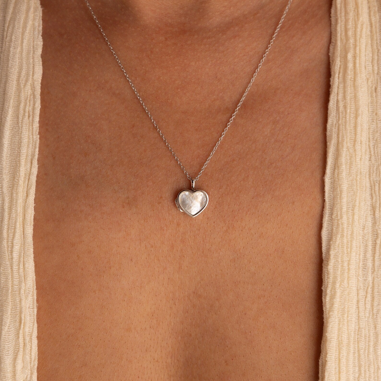 The Pearl Heart Locket Necklace, a nostalgic silver pendant, rests on the bare chest and is paired with a cream-colored garment.