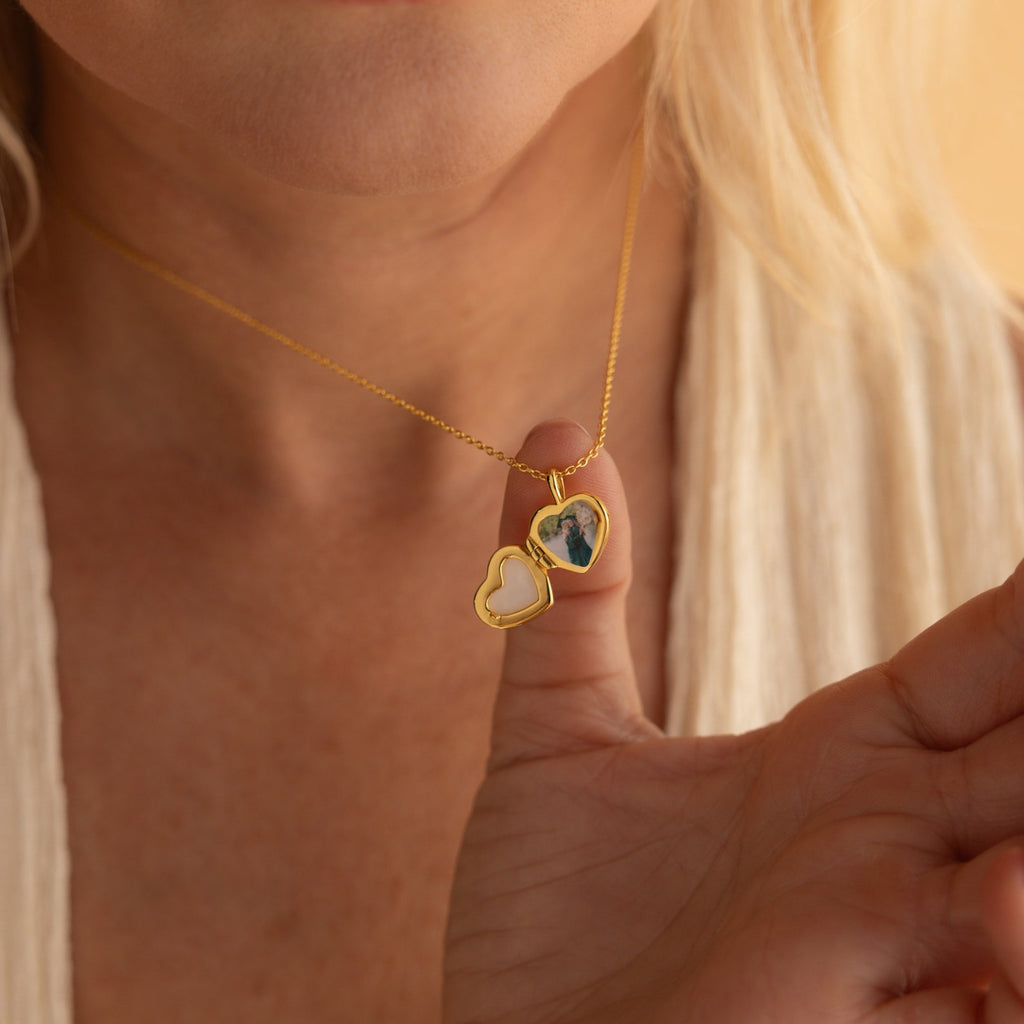 A woman gently holds a Pearl Heart Locket Necklace, featuring a nostalgic heart design and a cherished photo inside, close to her lips.