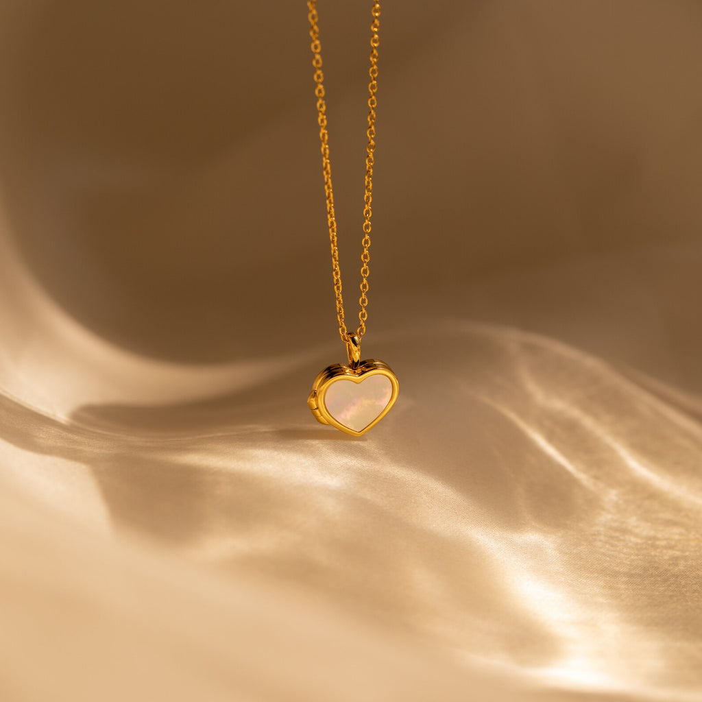 The Pearl Heart Locket Necklace features a gold chain with a Mother of Pearl heart locket pendant, showcased on a soft beige background—a timeless piece of nostalgic jewelry.