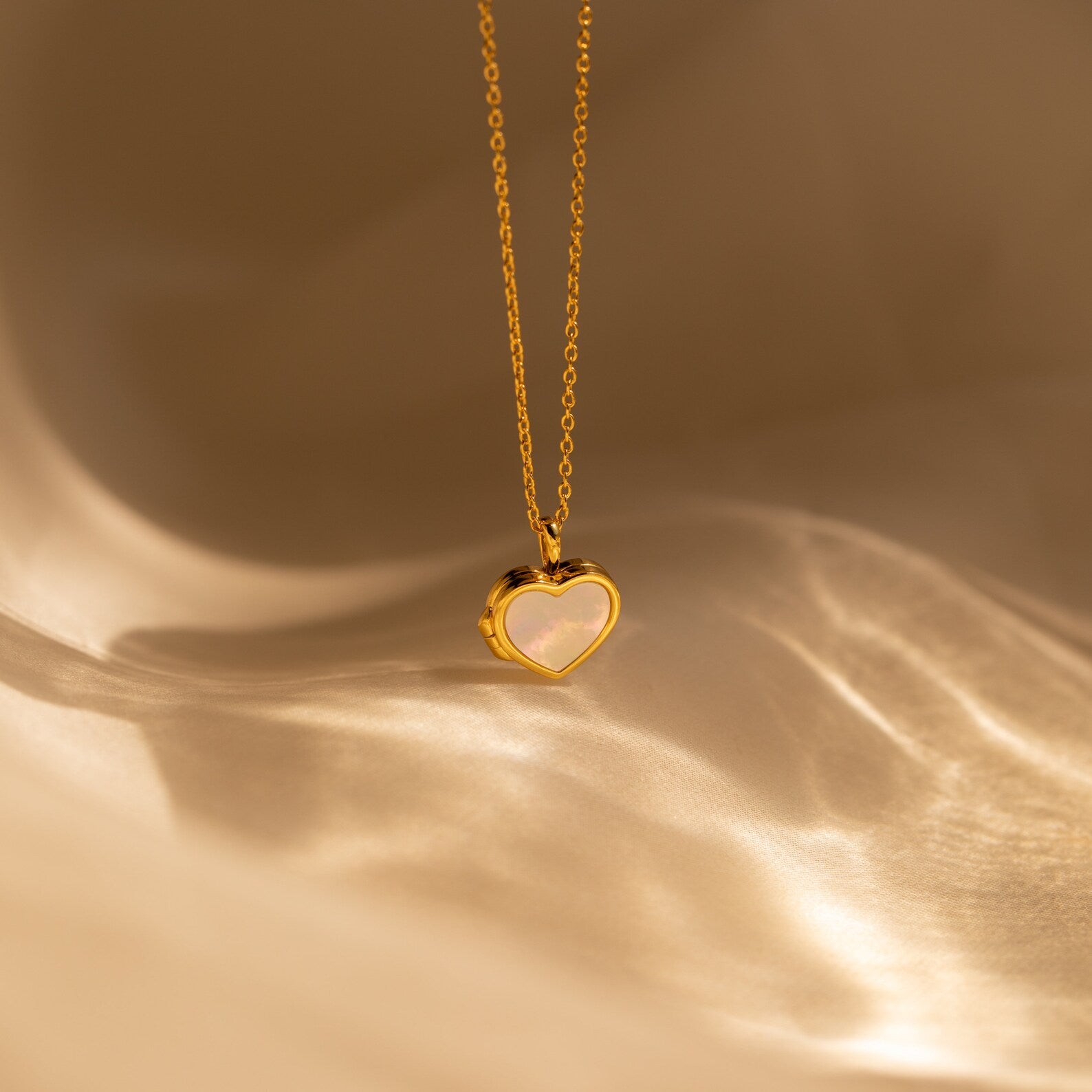 The Pearl Heart Locket Necklace features a gold chain with a Mother of Pearl heart locket pendant, showcased on a soft beige background—a timeless piece of nostalgic jewelry.