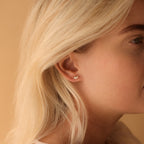 Close-up of a woman's ear adorned with playful earrings—tiny heart-shaped studs—against a soft beige background, perfect for fans of floral jewelry like Opal Marquise Studs.