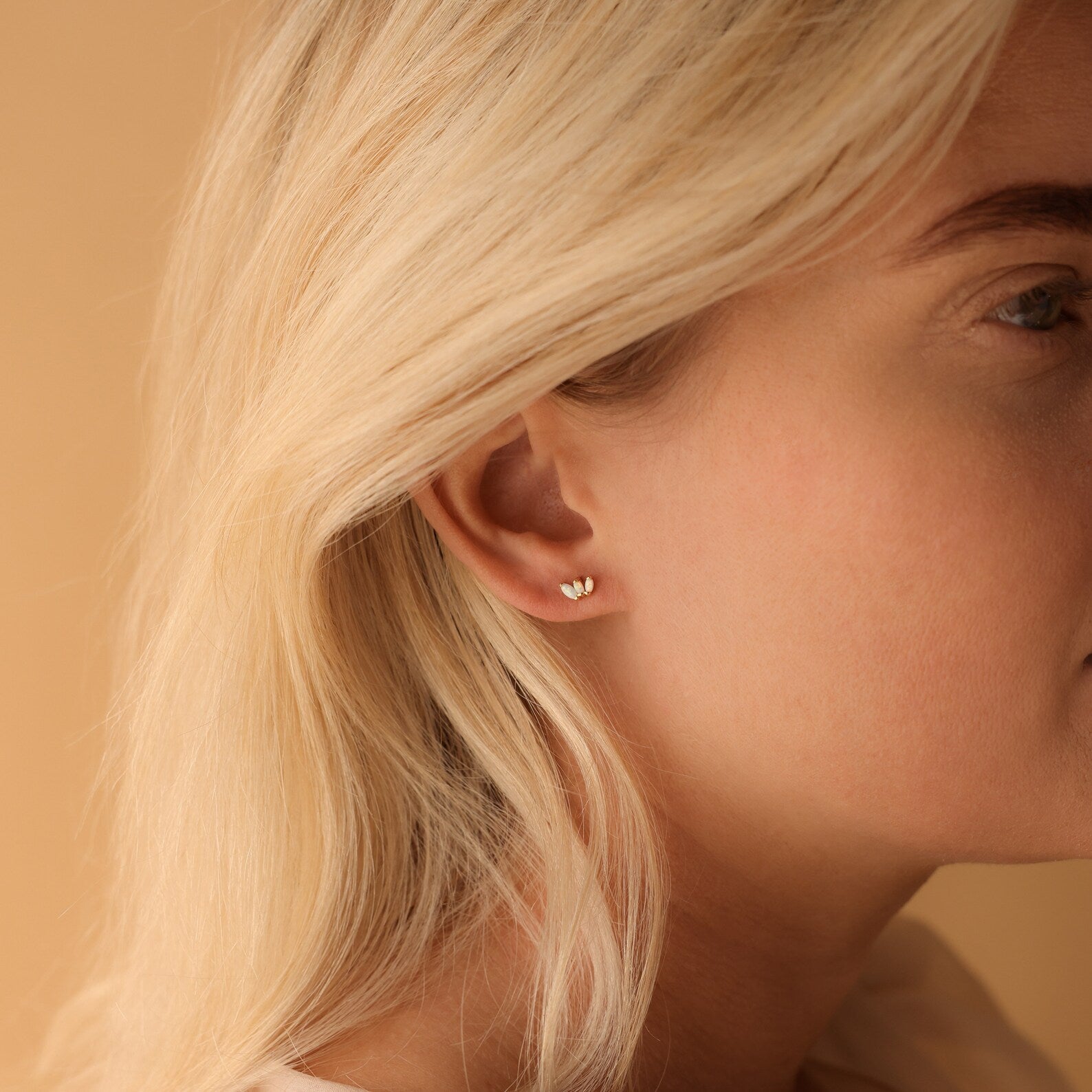 Close-up of a woman's ear adorned with playful earrings—tiny heart-shaped studs—against a soft beige background, perfect for fans of floral jewelry like Opal Marquise Studs.