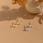 Two pairs of Opal Marquise Studs, one gold and one silver, are displayed on a beige surface near a white vase and dried flowers—a charming showcase of floral-inspired jewelry.