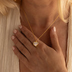 A woman with pale nails wears a gold Pearl Heart Locket Necklace and rests her hand on her chest.