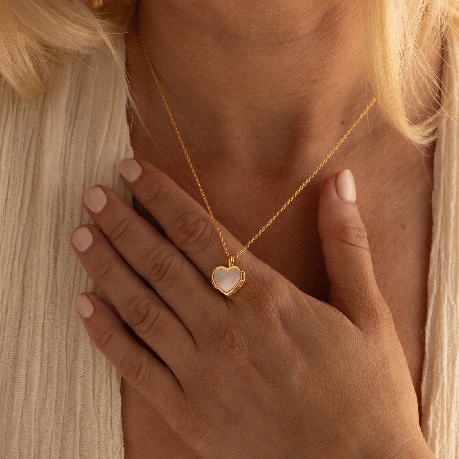 A woman with pale nails wears a gold Pearl Heart Locket Necklace and rests her hand on her chest.