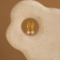 Pink Opal Drop Huggies, gold huggie hoop earrings featuring small pink opal gemstone dangles, shown on a textured beige stone background.