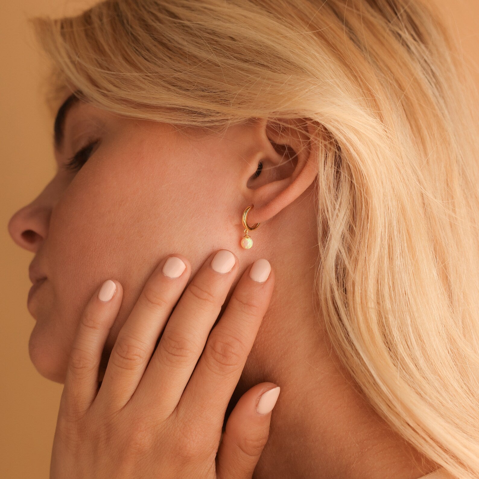 A woman with blonde hair touches her ear, showcasing a gold huggie hoop earring called Pink Opal Drop Huggies.