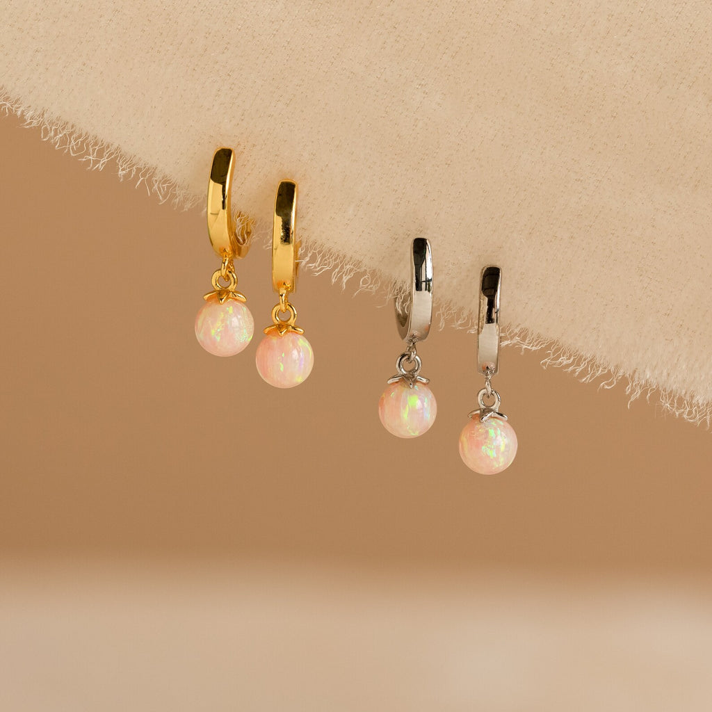 The Pink Opal Drop Huggies, featuring one gold and one silver pair, are adorned with pink opal charms and displayed on a beige fabric background.