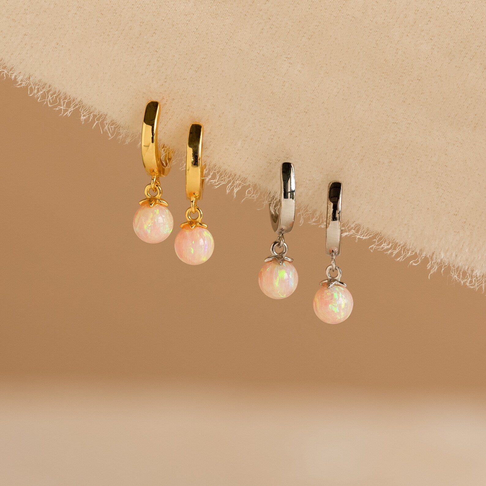 The Pink Opal Drop Huggies, featuring one gold and one silver pair, are adorned with pink opal charms and displayed on a beige fabric background.