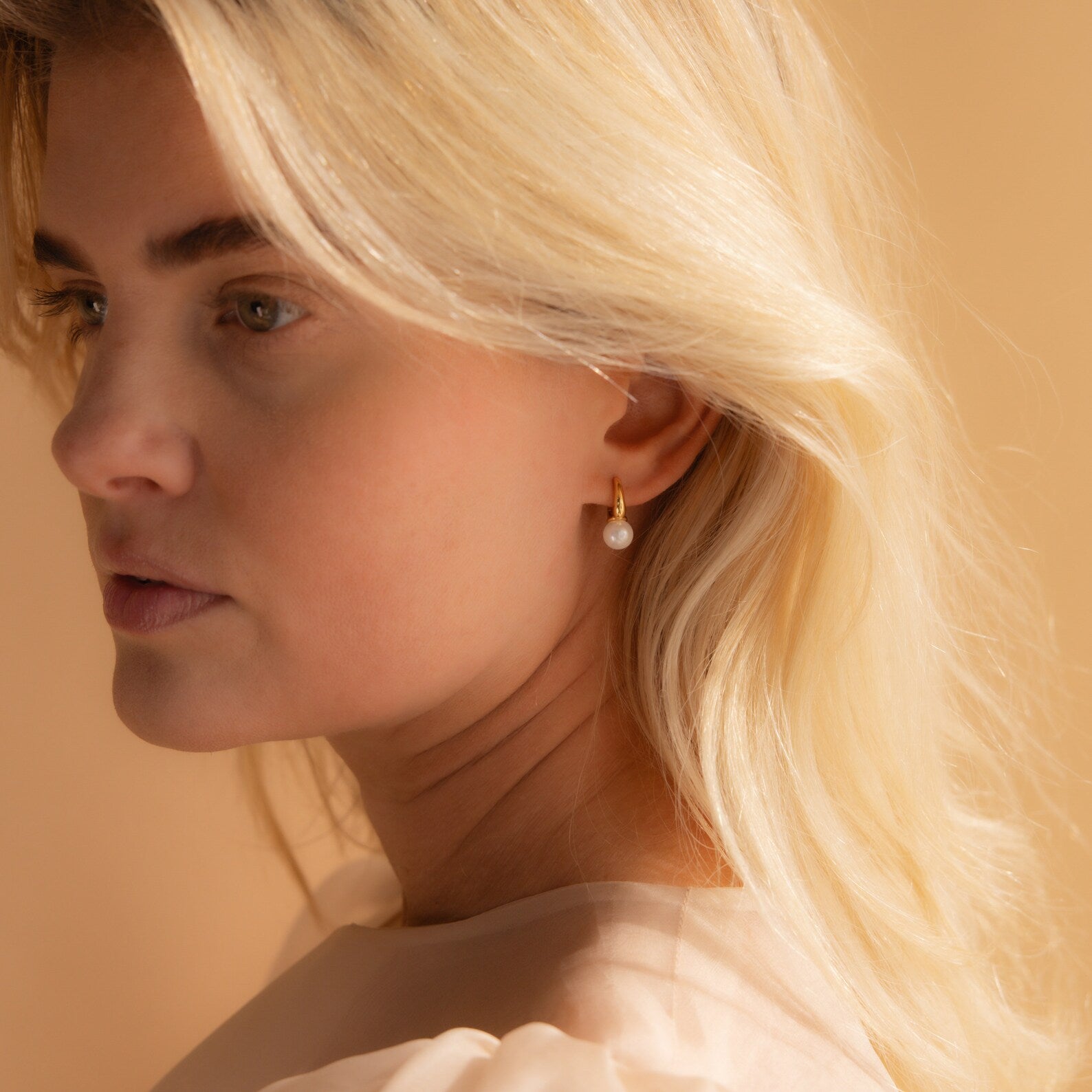  three-quarter portrait of a blonde woman wearing a single gold Pearl Drop Huggie, her gaze directed softly to the side.