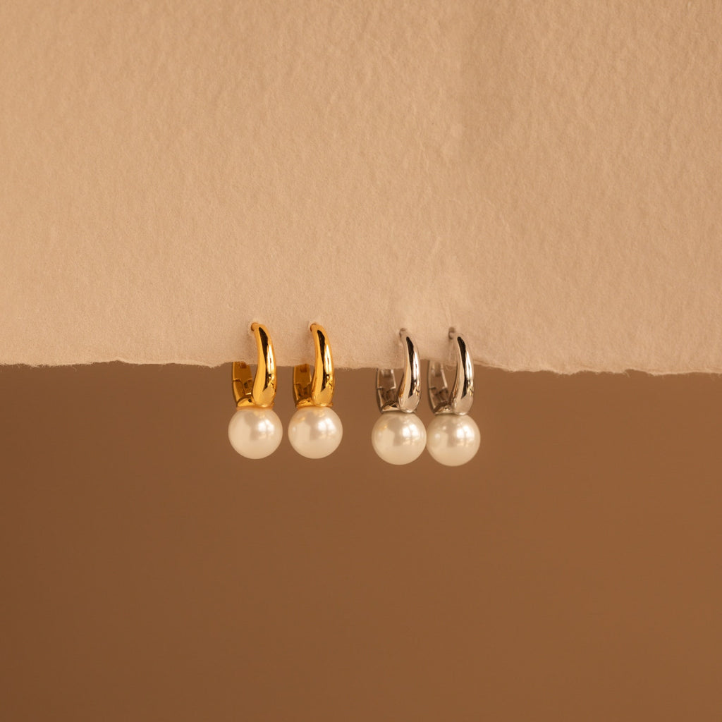 A pair of elegant Pearl Drop Huggie earrings is displayed against a textured, cream-colored paper backdrop. The design includes two sets of huggie hoops—one in shiny gold and the other in sleek silver—each holding a single lustrous round white pearl that dangles delicately beneath the hoop.