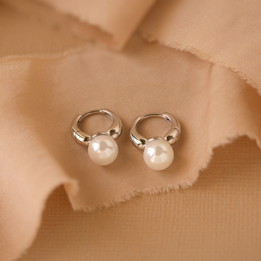 A close-up image showcasing a pair of silver-tone huggie hoop earrings, each adorned with a single lustrous white pearl.The hoops have a smooth, high-polished finish, reflecting light and their surroundings with a mirror-like gleam