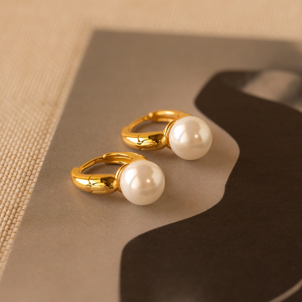 A close-up image of a pair of gold-tone huggie earrings, each adorned with a single large, luminous white pearl. The polished gold finish of the hoops gleams under soft lighting, emphasizing their smooth curves and the high-shine luster of the pearls. Subtle reflections on the pearls showcase their perfect spherical shape and creamy nacre finish