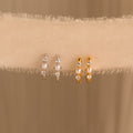 Two pairs of pearl and diamond huggies—one in gold, one in silver—displayed on a fabric ribbon for contrast.