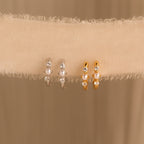 Two pairs of pearl and diamond huggies—one in gold, one in silver—displayed on a fabric ribbon for contrast.