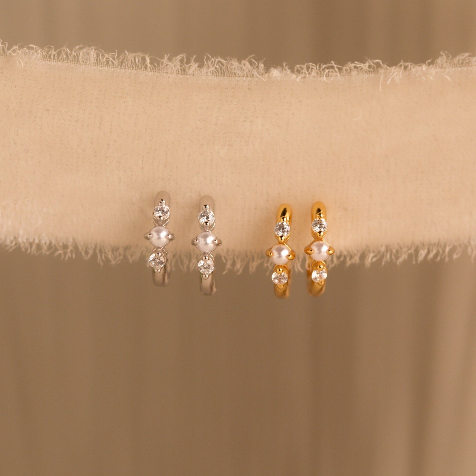 Two pairs of pearl and diamond huggies—one in gold, one in silver—displayed on a fabric ribbon for contrast.
