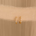 A pair of gold huggie earrings with pearl and diamond accents hanging against a soft cream fabric backdrop.
