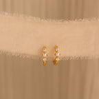 A pair of gold huggie earrings with pearl and diamond accents hanging against a soft cream fabric backdrop.