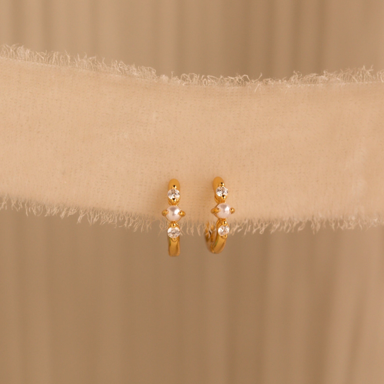 A pair of gold huggie earrings with pearl and diamond accents hanging against a soft cream fabric backdrop.