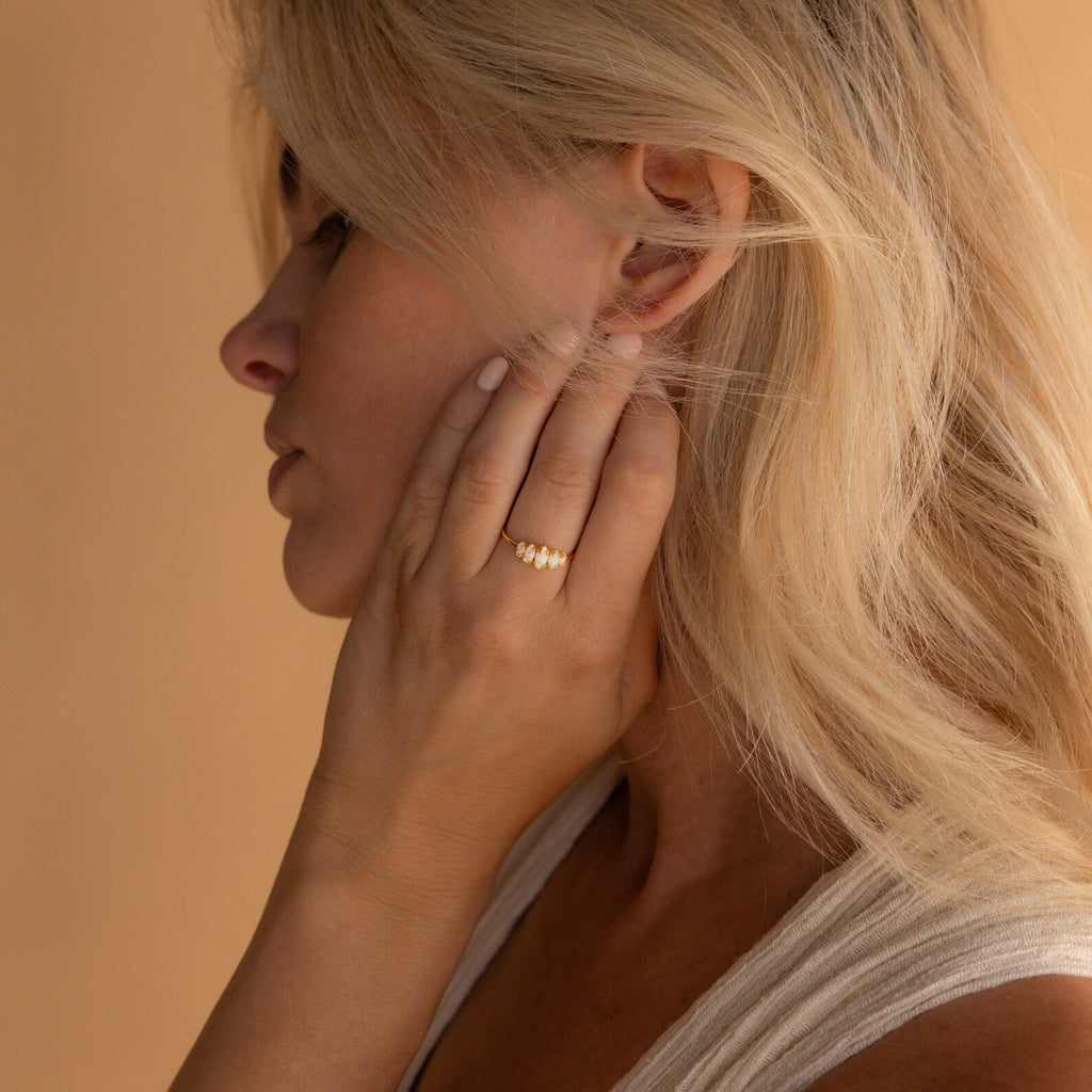 A woman with blonde hair touches her ear, highlighting a Marquise Opal Statement Ring and a sleeveless light top, radiating classic style.