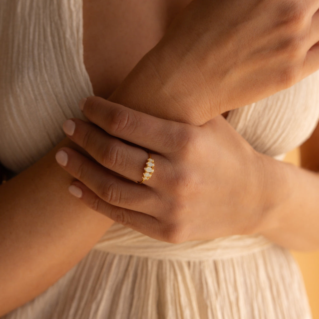 A woman in a cream dress wears the Marquise Opal Statement Ring, its opal gem radiating vintage allure and classic elegance as her hands are gently clasped over her waist.