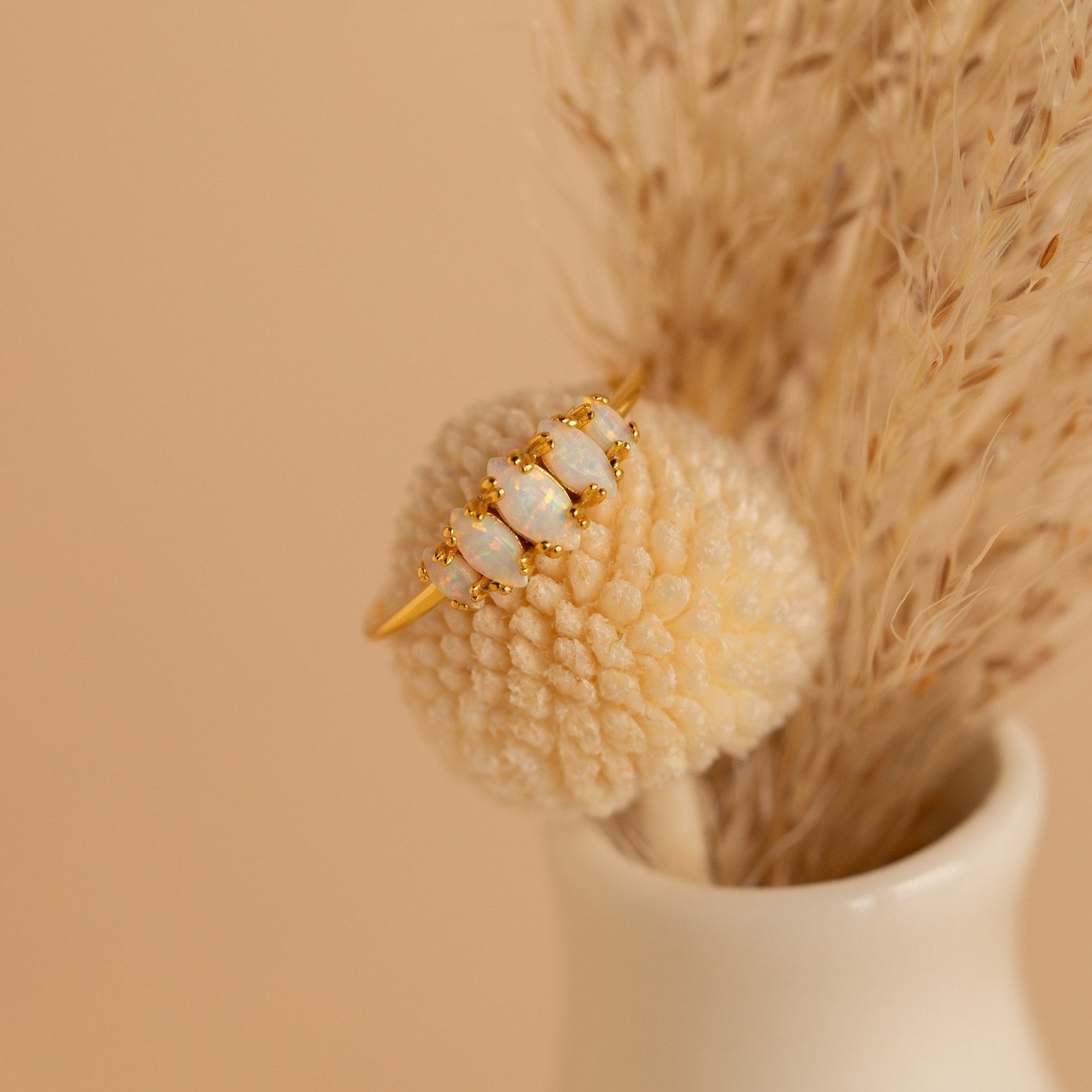 Exuding vintage charm, the Marquise Opal Statement Ring with four white stones is displayed on a textured cream ball, complemented by dried grasses in a vase.