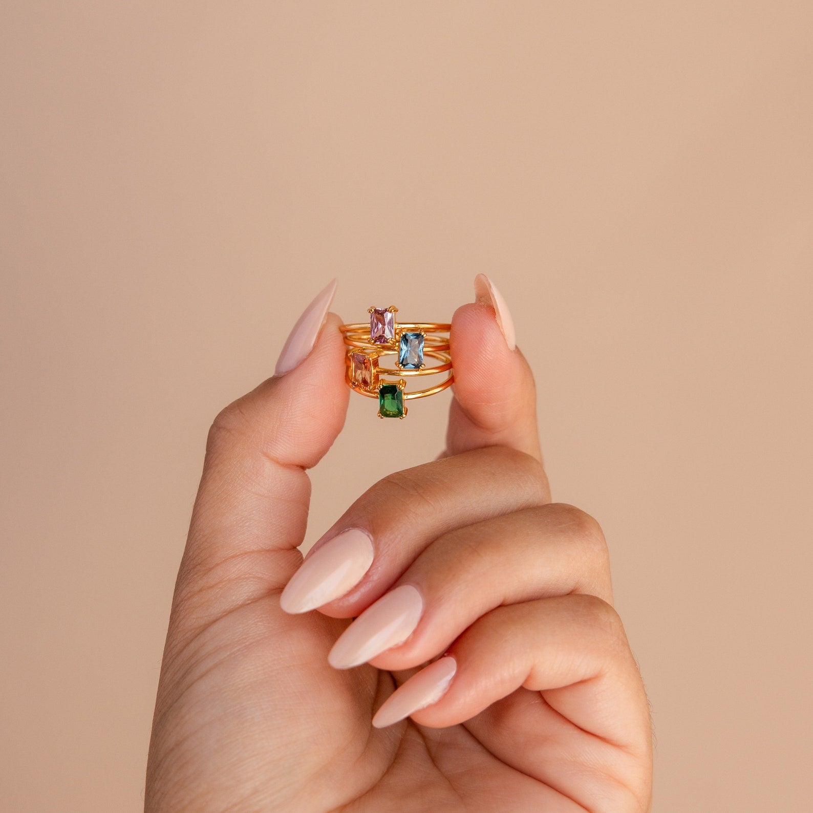 A hand with nude nails displays three gemstone rings, featuring the Emerald Cut Solitaire Rings collection, against a beige background.
