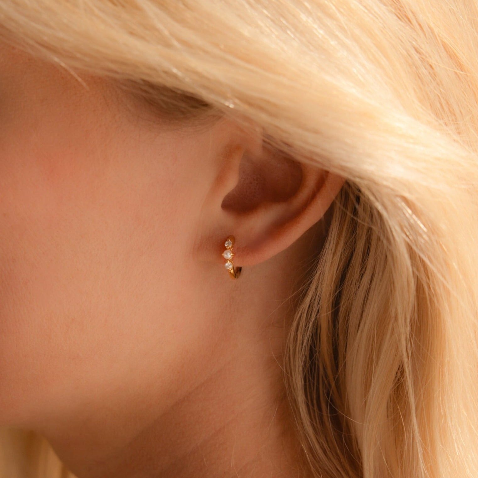 A close-up shot of a woman's right ear adorned with a dainty gold huggie earring, styled with alternating round-cut diamonds and tiny freshwater pearls. The three stones—diamond, pearl, diamond—are bezel-set into a polished gold hoop that gently hugs the earlobe.