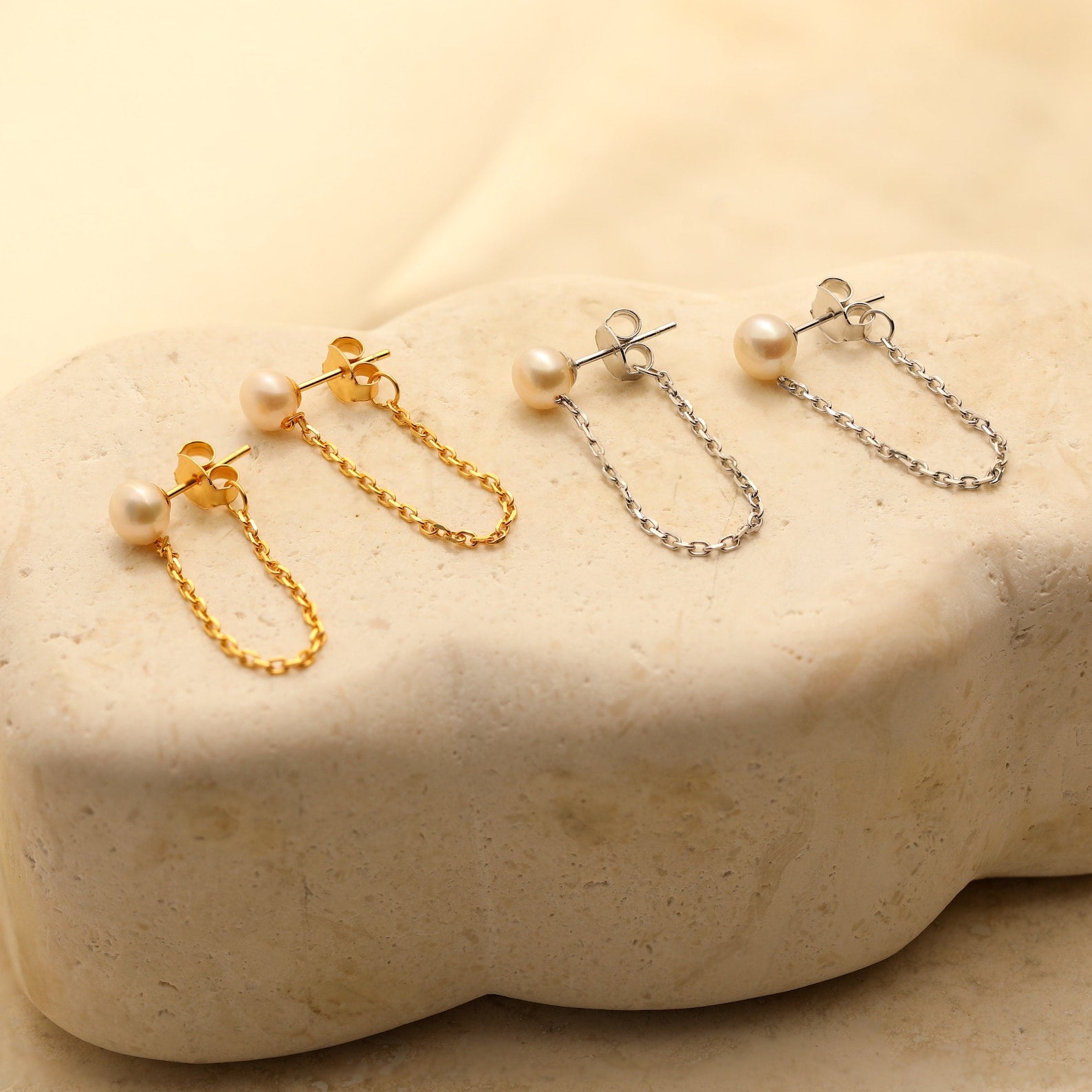 Two pairs of Pearl Chain Studs, one in gold and one in silver, are displayed on a beige stone surface.