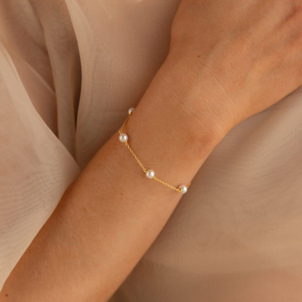 The Pearl Station Bracelet features luminous pearls on a gold chain, shown elegantly draped on a woman's wrist against beige fabric—perfect for modern pearl jewelry lovers.