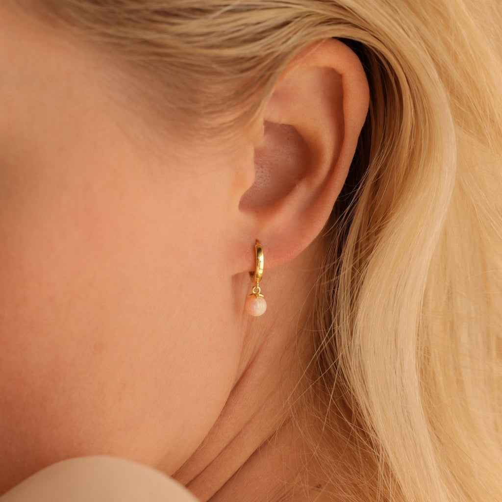Close-up of a woman's ear wearing Pink Opal Drop Huggies, featuring huggie hoops adorned with a delicate pink opal charm.