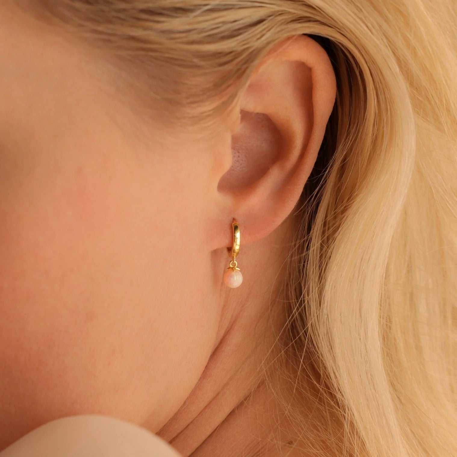 Close-up of a woman's ear wearing the Opal Drop Earrings Set—small gold hoops with light pink opal dangles, perfect for layering. Blonde hair visible.