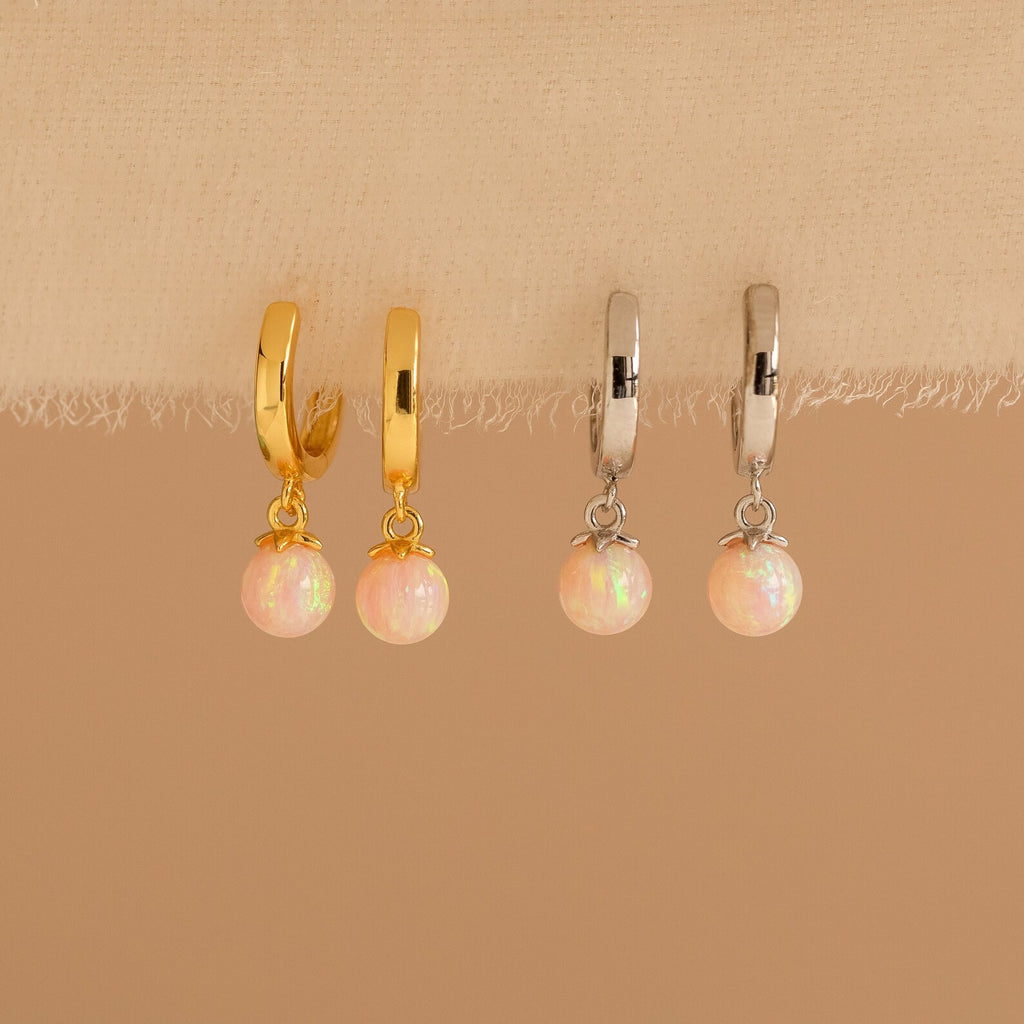 Two pairs of Pink Opal Drop Huggies, one gold and one silver, are displayed on fabric. Each features a delicate pink opal-like charm that adds a subtle sparkle to any look.