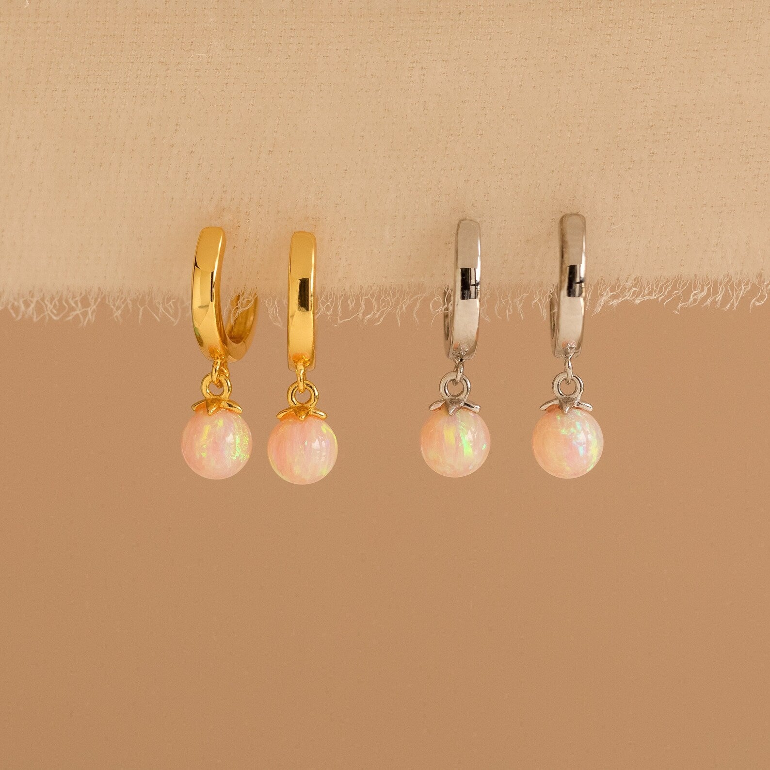 Two pairs of Pink Opal Drop Huggies, one gold and one silver, are displayed on fabric. Each features a delicate pink opal-like charm that adds a subtle sparkle to any look.
