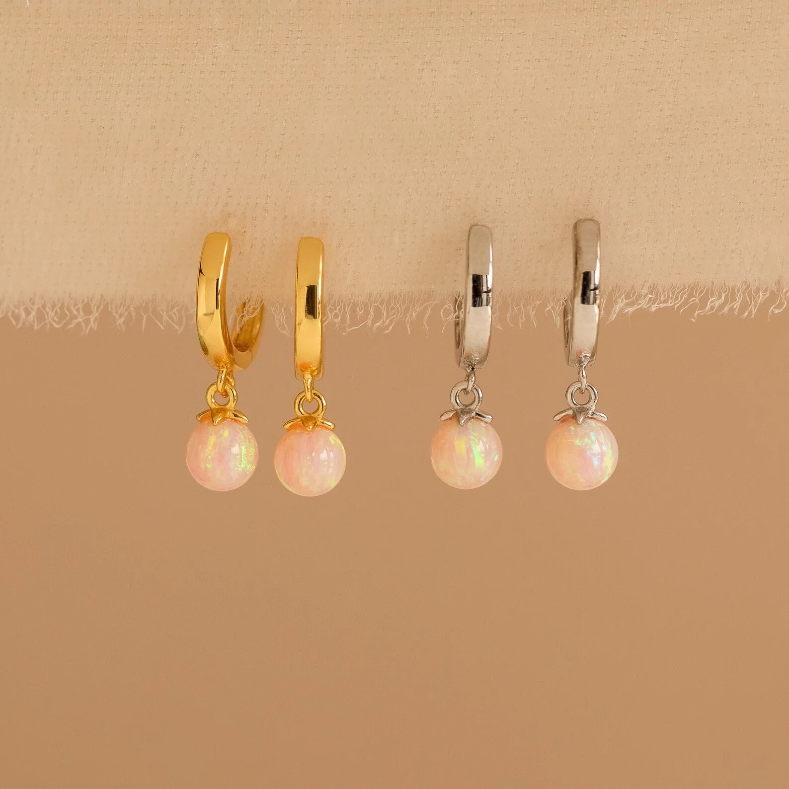 The Opal Drop Earrings Set features two pairs of hoop earrings—one gold and one silver—with opal dangles, perfect for layering your jewelry on a beige fabric display.
