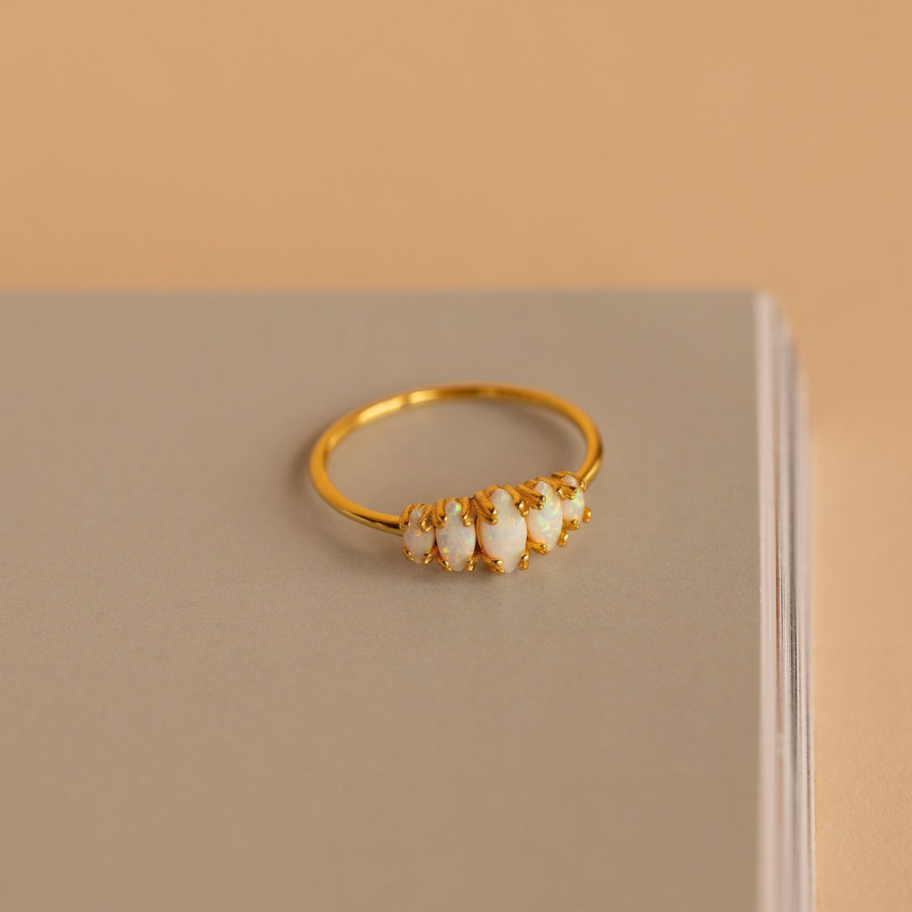 The Marquise Opal Statement Ring features five white opals, shown on a closed beige book against a soft beige background, radiating vintage allure and classic style.
