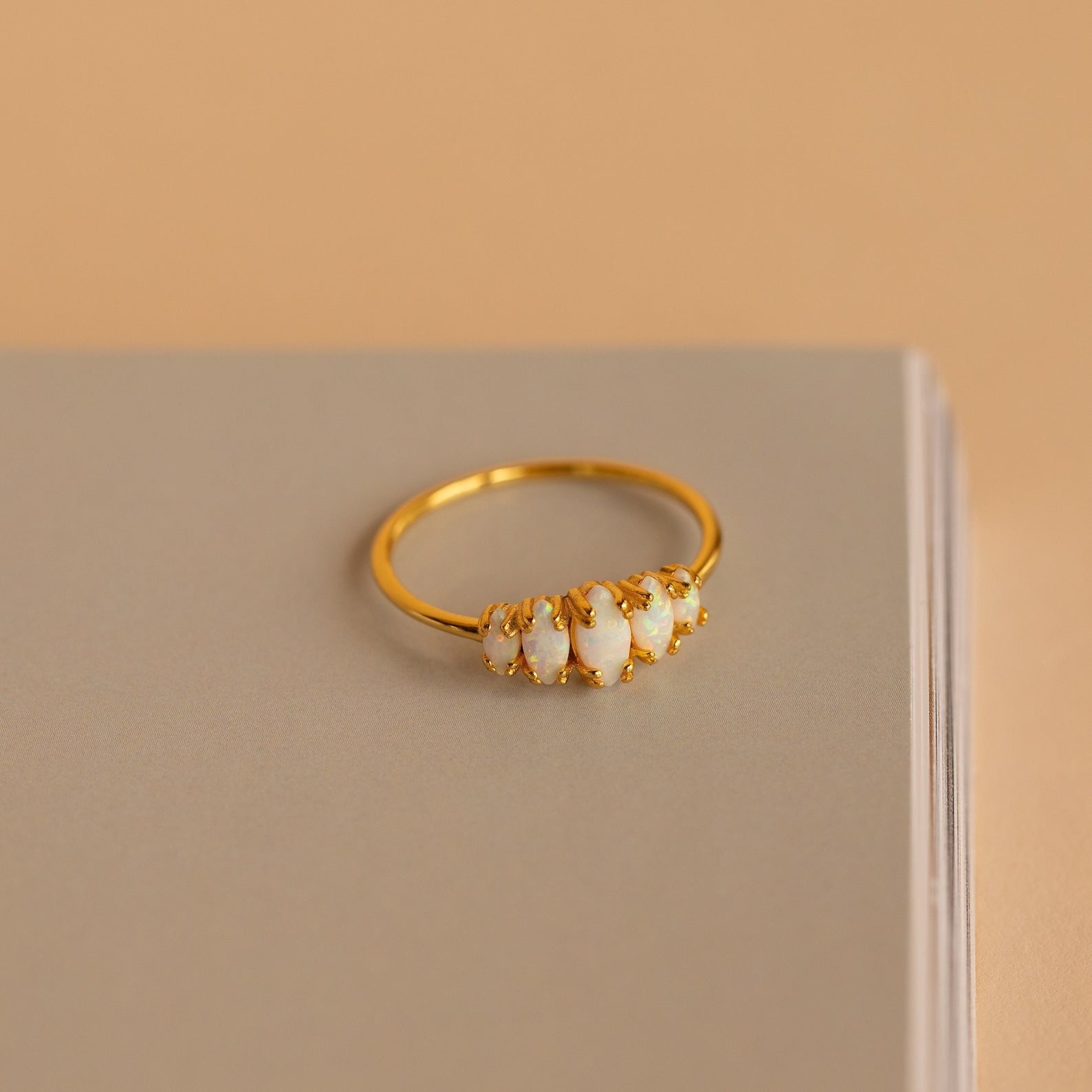 The Marquise Opal Statement Ring features five white opals, shown on a closed beige book against a soft beige background, radiating vintage allure and classic style.
