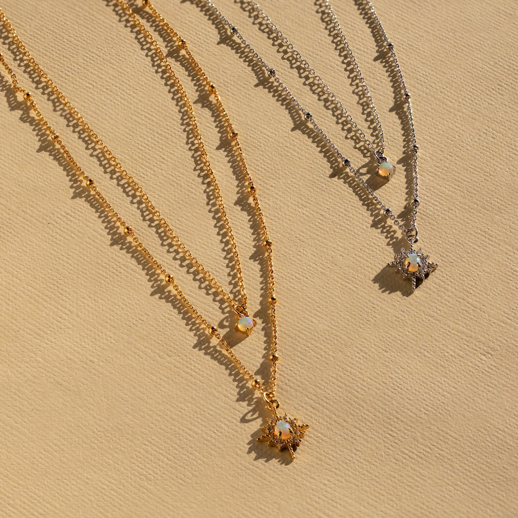 The Opal Star Duo Necklace features two layered necklaces, one gold and one silver, each with a starburst pendant, elegantly displayed on a beige background.