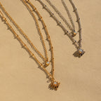 The Opal Star Duo Necklace features two layered necklaces, one gold and one silver, each with a starburst pendant, elegantly displayed on a beige background.