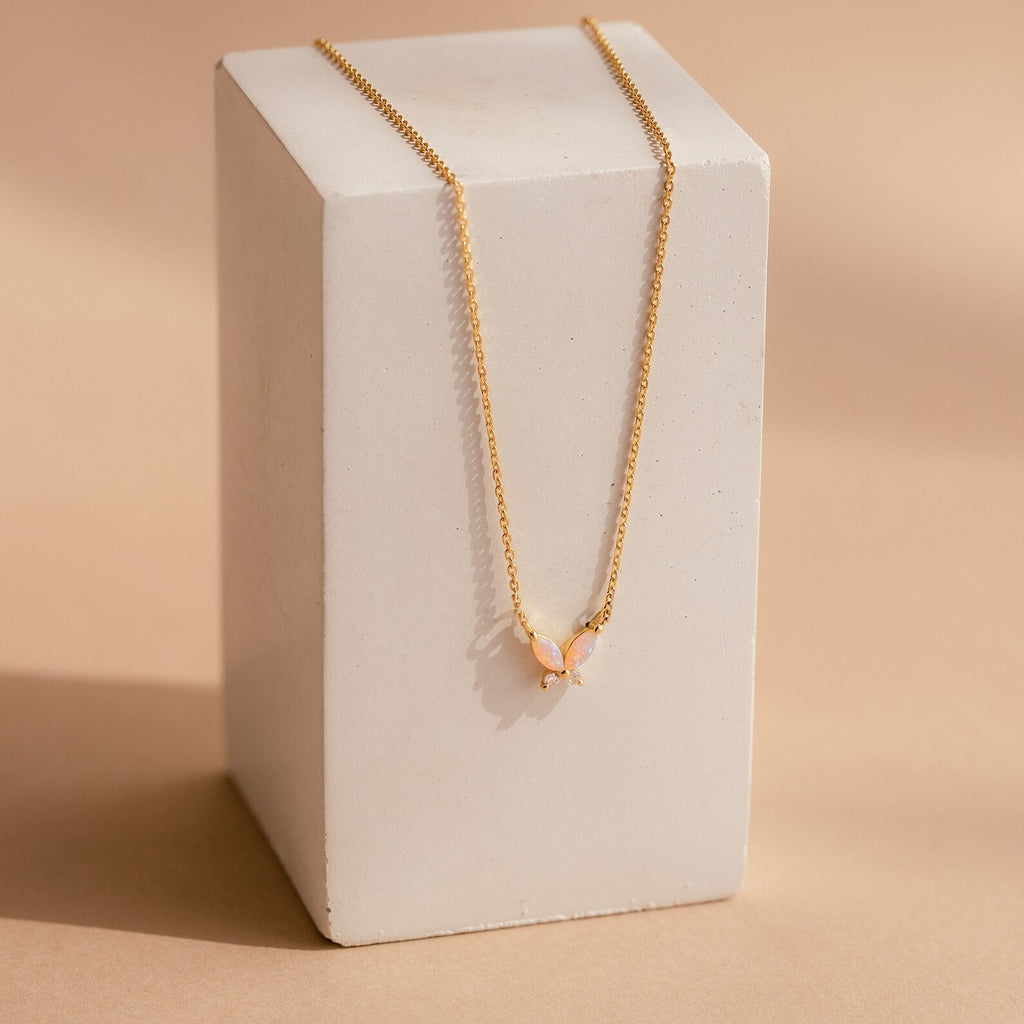 The Pink Opal Butterfly Necklace features a gold chain with a delicate pink opal butterfly pendant, beautifully displayed on a rectangular white block for an elegant touch.